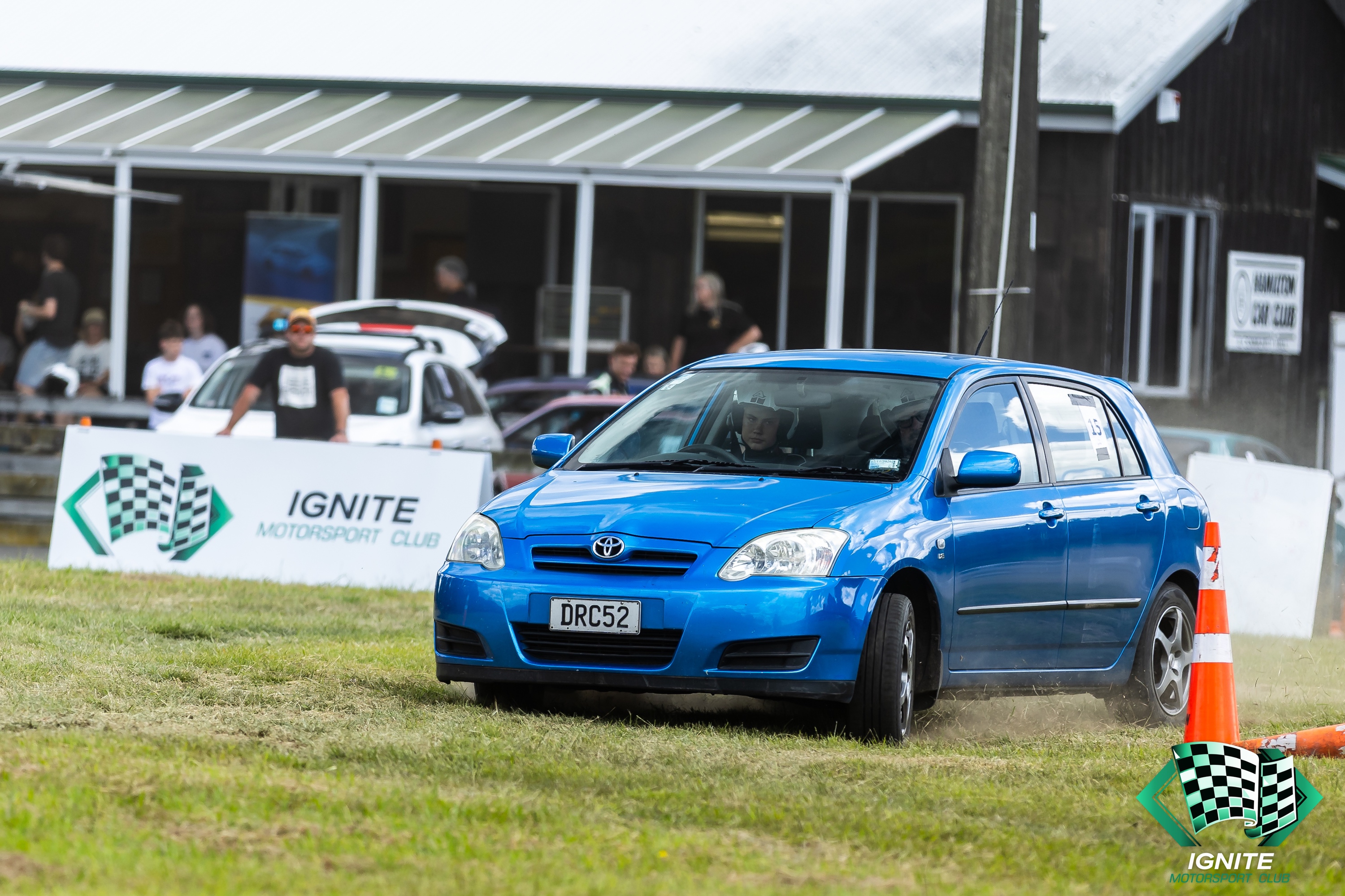 A young driver in a blue hatchback rounding a cone at an Ignite autocross